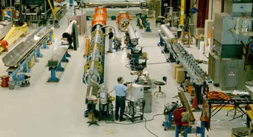 The magnet test area at Fermilab with the first quadrupole-corrector magnet assembly, consisting of two Fermilab quadrupoles and a CERN correction coil. Denny Gaw (foreground) and Jan Szal perform a leak check before the assembly is inserted into its cryostat vessel.